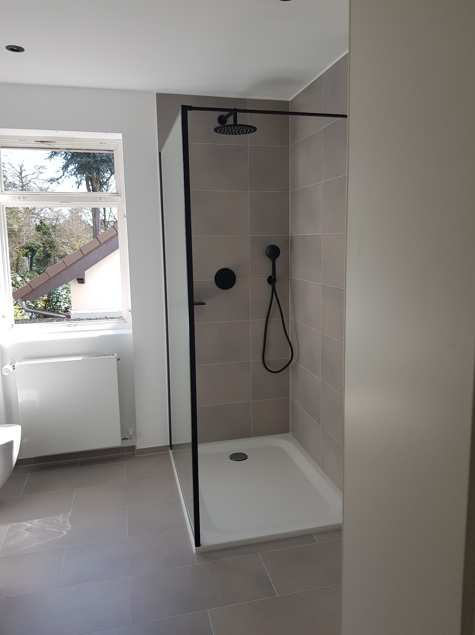 Nowoczesna kabina prysznicowa z czarnymi detalami, szare płytki na ścianach i podłodze, widok z boku, okno z widokiem na zewnątrz, minimalistyczny design.