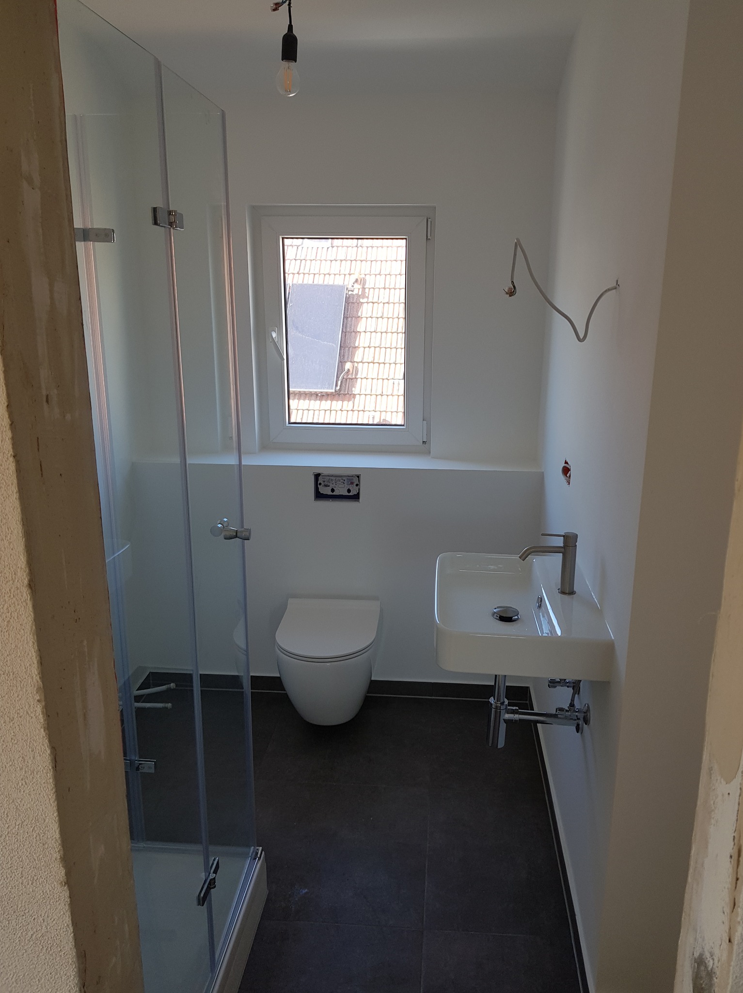 Nowoczesna łazienka w trakcie wykończenia: biała armatura (umywalka, toaleta), szklana kabina prysznicowa, ciemna podłoga. Widoczne niedokończone elementy instalacji.