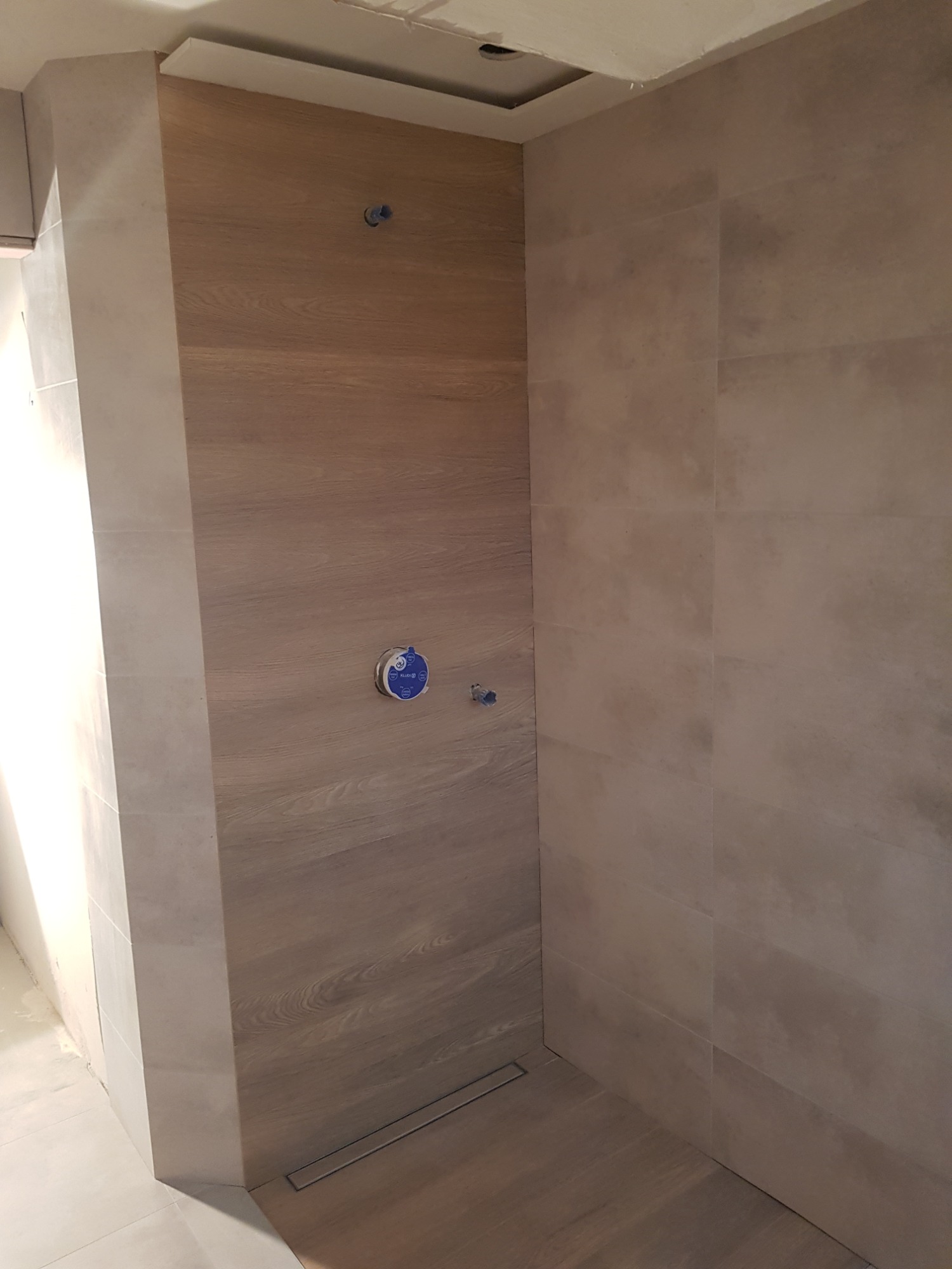 Wykończona kabina prysznicowa z drewnopodobnymi płytkami na jednej ścianie i beżowymi na pozostałych, widoczne odpływ liniowy i przygotowane punkty instalacyjne