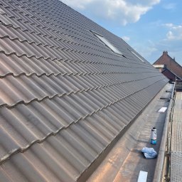 SHARKO SPÓŁKA Z OGRANICZONĄ ODPOWIEDZIALNOŚCIĄ - Ciemna dachówka ceramiczna na skośnym dachu z oknami połaciowymi, widok z rusztowania. Obróbki blacharskie wzdłuż krawędzi dachu. Na pierwszym planie narzędzia.