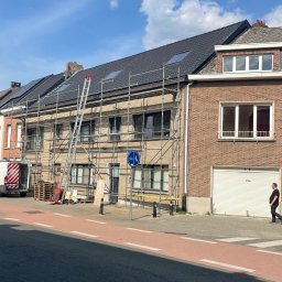 SHARKO SPÓŁKA Z OGRANICZONĄ ODPOWIEDZIALNOŚCIĄ - Dom w rusztowaniach z nowym, ciemnym dachem i oknami dachowymi. Na chodniku przechodzień, a obok zaparkowany van. Słoneczny dzień.