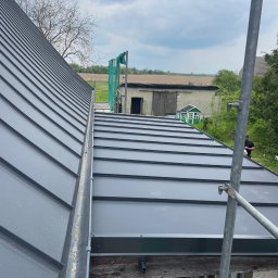 SHARKO SPÓŁKA Z OGRANICZONĄ ODPOWIEDZIALNOŚCIĄ - Szary dach z blachy na rąbek stojący, widok z rusztowania. W tle budynek gospodarczy i zielony domek dla dzieci. Nowoczesne krycie dachu.