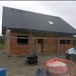 SHARKO SPÓŁKA Z OGRANICZONĄ ODPOWIEDZIALNOŚCIĄ - Budowa domu w trakcie: murowane ściany z pustaków, nowy dach z ciemnej dachówki, rusztowania. Na pierwszym planie betoniarka i paleta z dachówką.