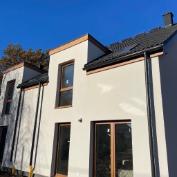 SHARKO SPÓŁKA Z OGRANICZONĄ ODPOWIEDZIALNOŚCIĄ - Nowoczesny dom z ciemną dachówką i panelami solarnymi, jasną elewacją, drewnianymi ramami okiennymi i czarnymi rynnami w słoneczny dzień.