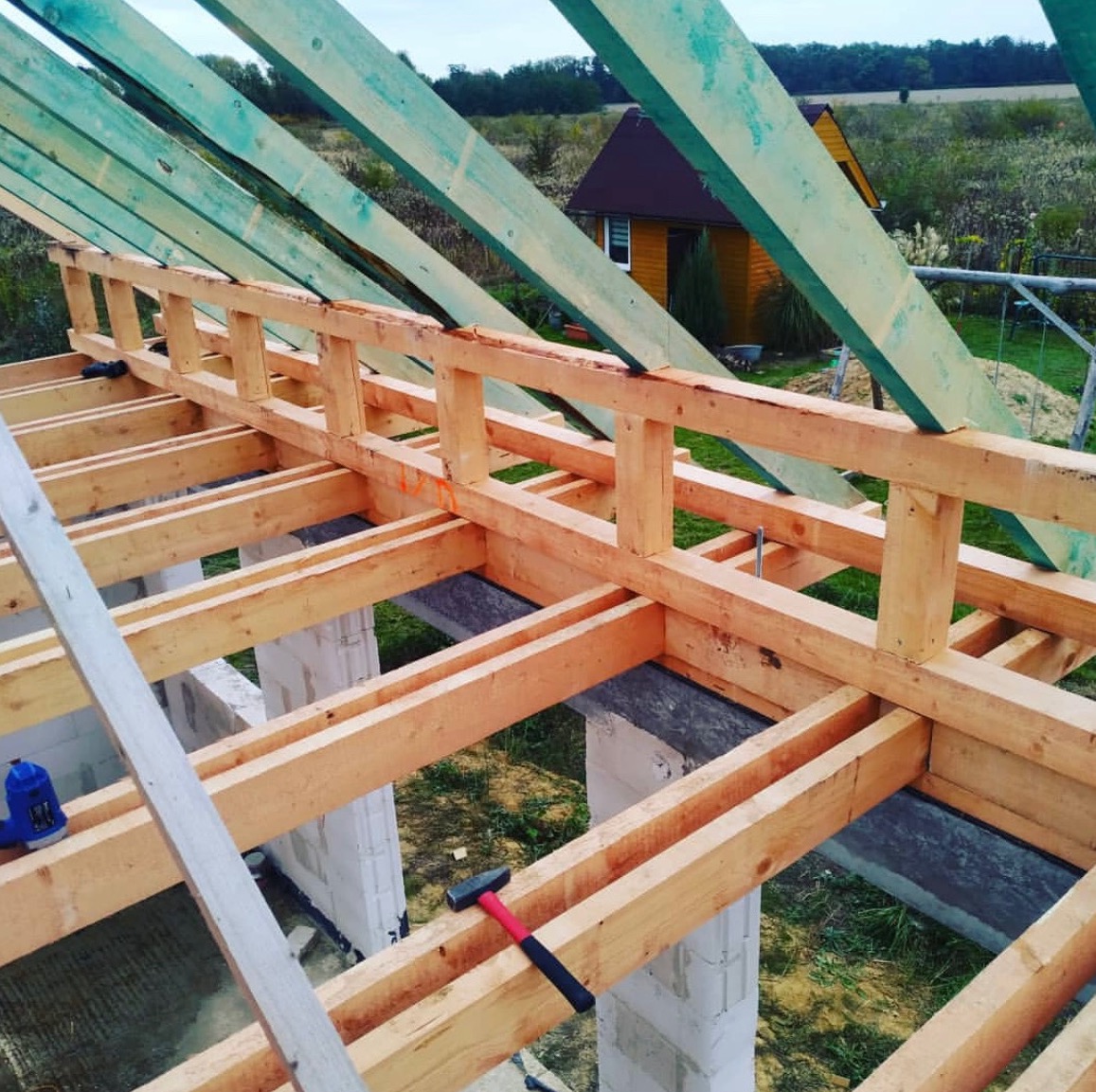 Wznoszenie drewnianej konstrukcji dachu z użyciem belek w kolorze naturalnego drewna i zielonych krokwi, na tle krajobrazu z małym domkiem i zielenią, widoczny młotek z czerwoną rączką.