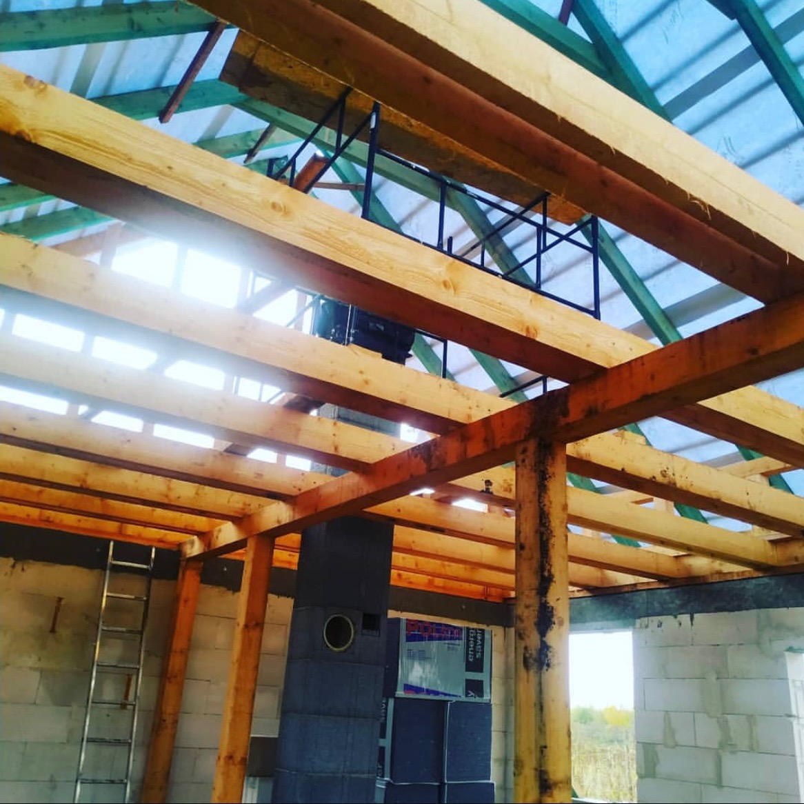 Wnętrze domu w trakcie budowy, widoczna drewniana konstrukcja więźby dachowej z elementami stalowymi, częściowo przykryta folią, komin z szarej cegły oraz drabina oparta o ścianę z białych bloczków.