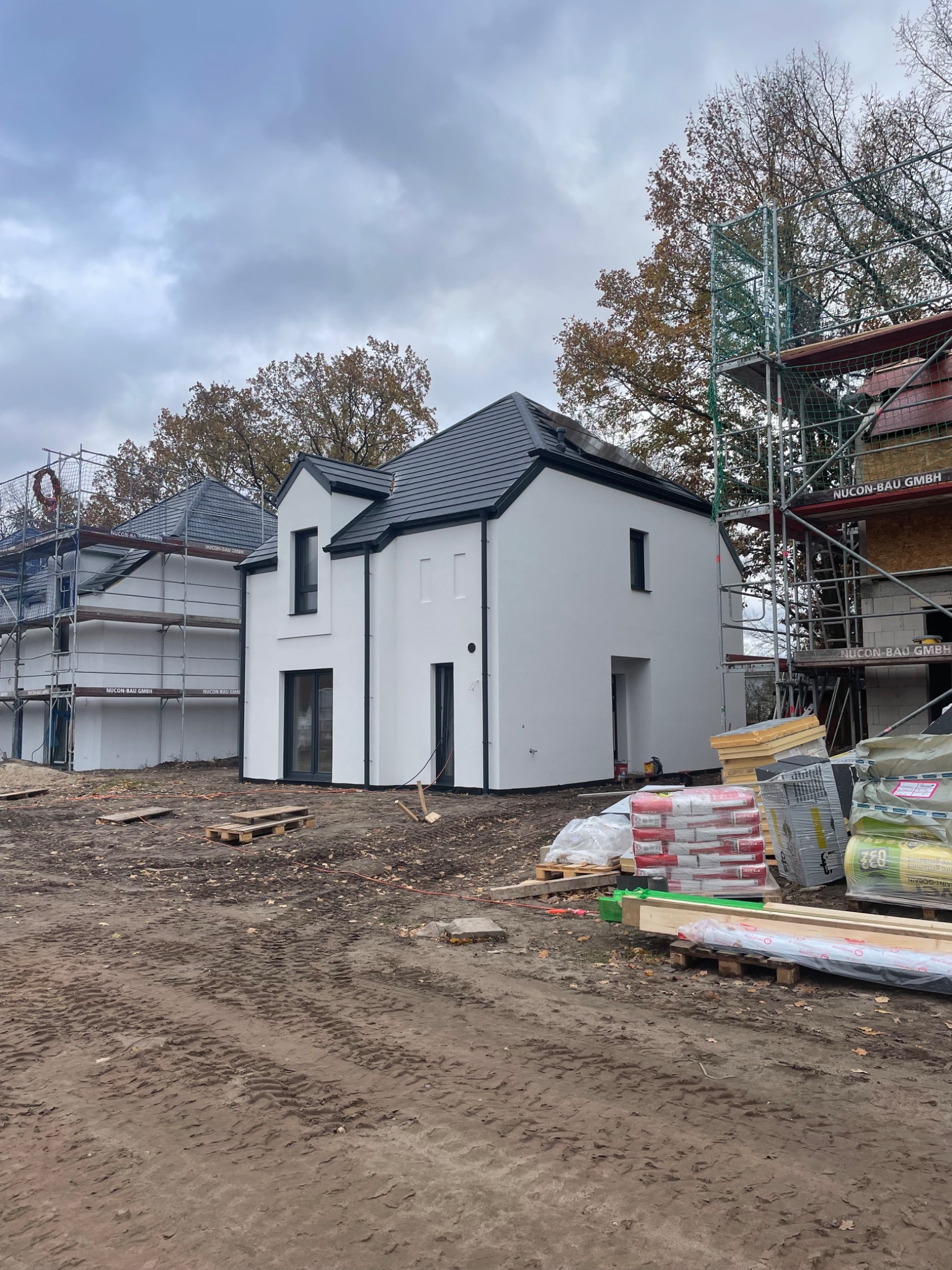 Dwa nowoczesne, białe domy w trakcie budowy, z czarnymi dachami i oknami, otoczone rusztowaniami i materiałami budowlanymi na placu budowy w pochmurny dzień.