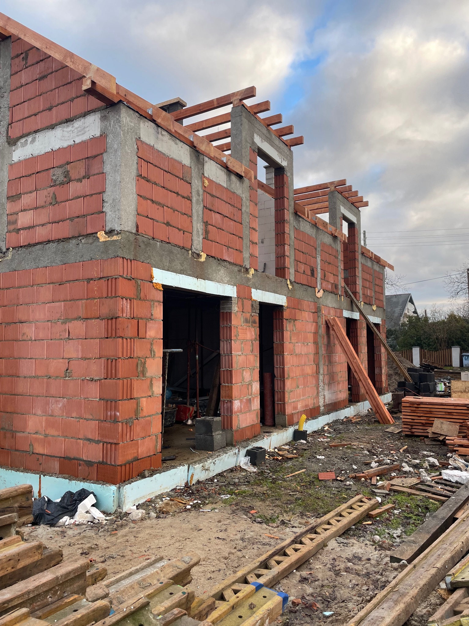 Budowa domu murowanego w Lübbenau/Spreewald na etapie wznoszenia ścian z czerwonej cegły i wylewania betonowych słupów, widoczne drewniane elementy konstrukcyjne i materiały budowlane na placu budowy.