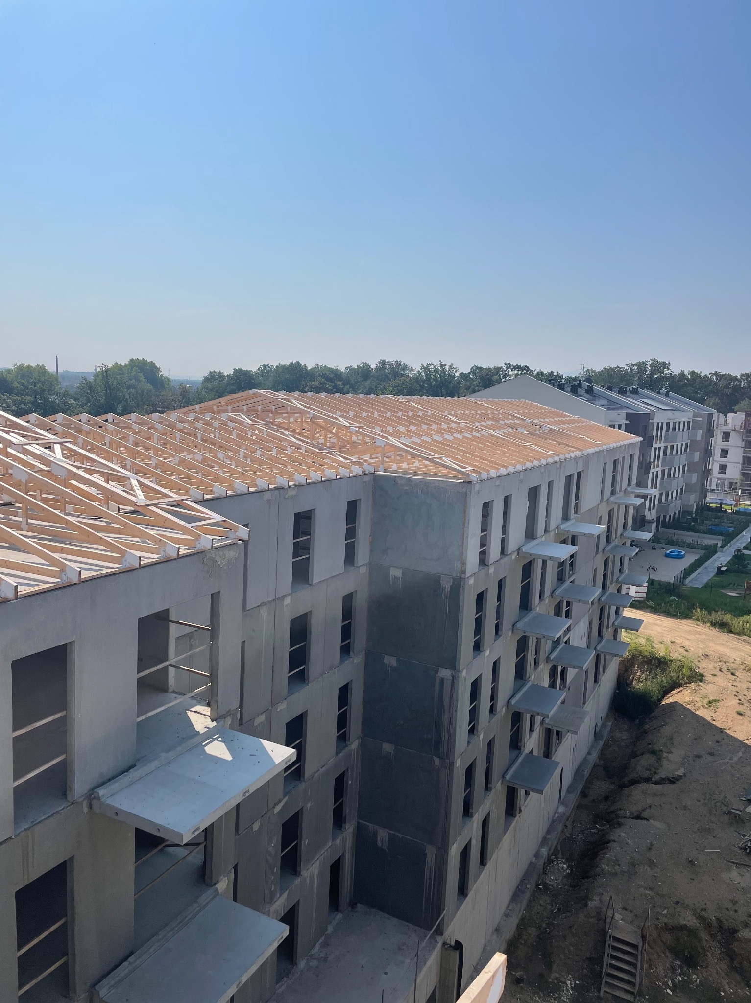 Wysoki budynek wielorodzinny w stanie surowym, widoczna konstrukcja drewniana dachu oraz betonowe balkony, w tle zieleń i inne budynki.