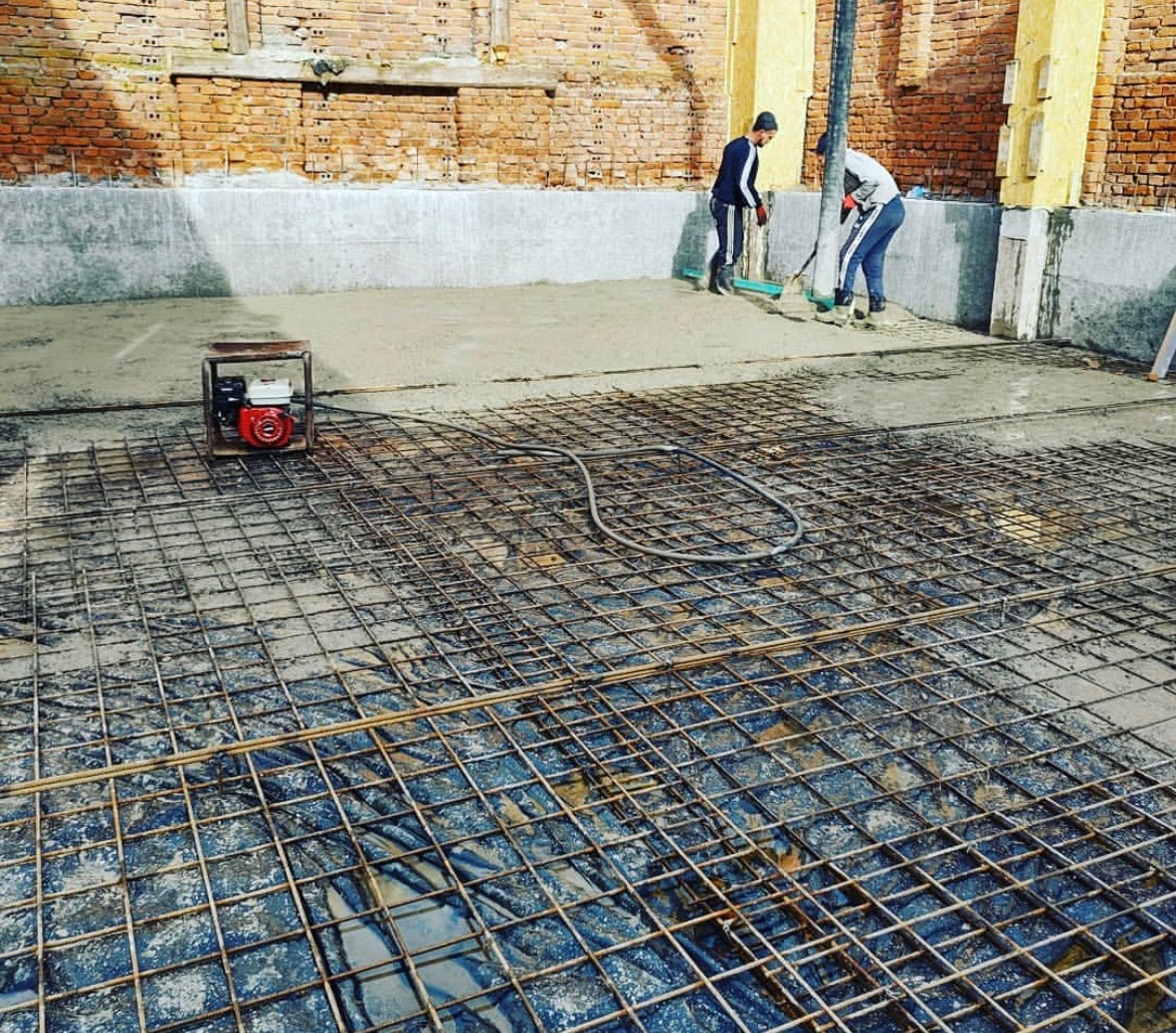 Zbrojenie fundamentów z wylanym betonem, widoczni dwaj pracownicy rozprowadzający beton. Na pierwszym planie siatka zbrojeniowa i agregat prądotwórczy.