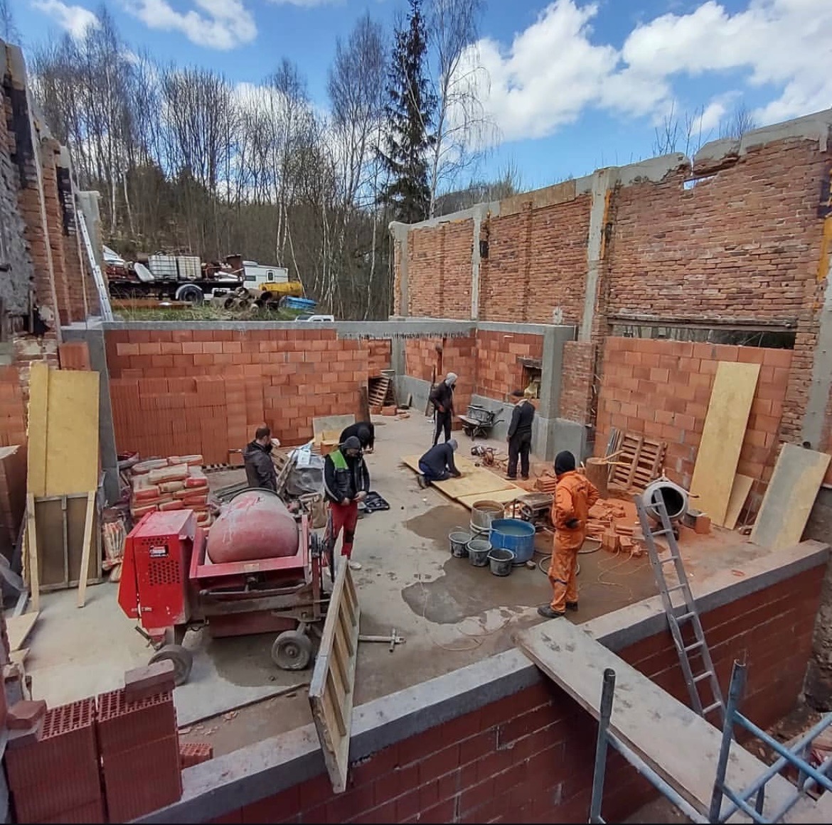 Budowa domu z cegły: ekipa murarzy w trakcie pracy, widoczna betoniarka, palety z cegłami i częściowo wzniesione ściany. W tle drzewa i błękitne niebo.