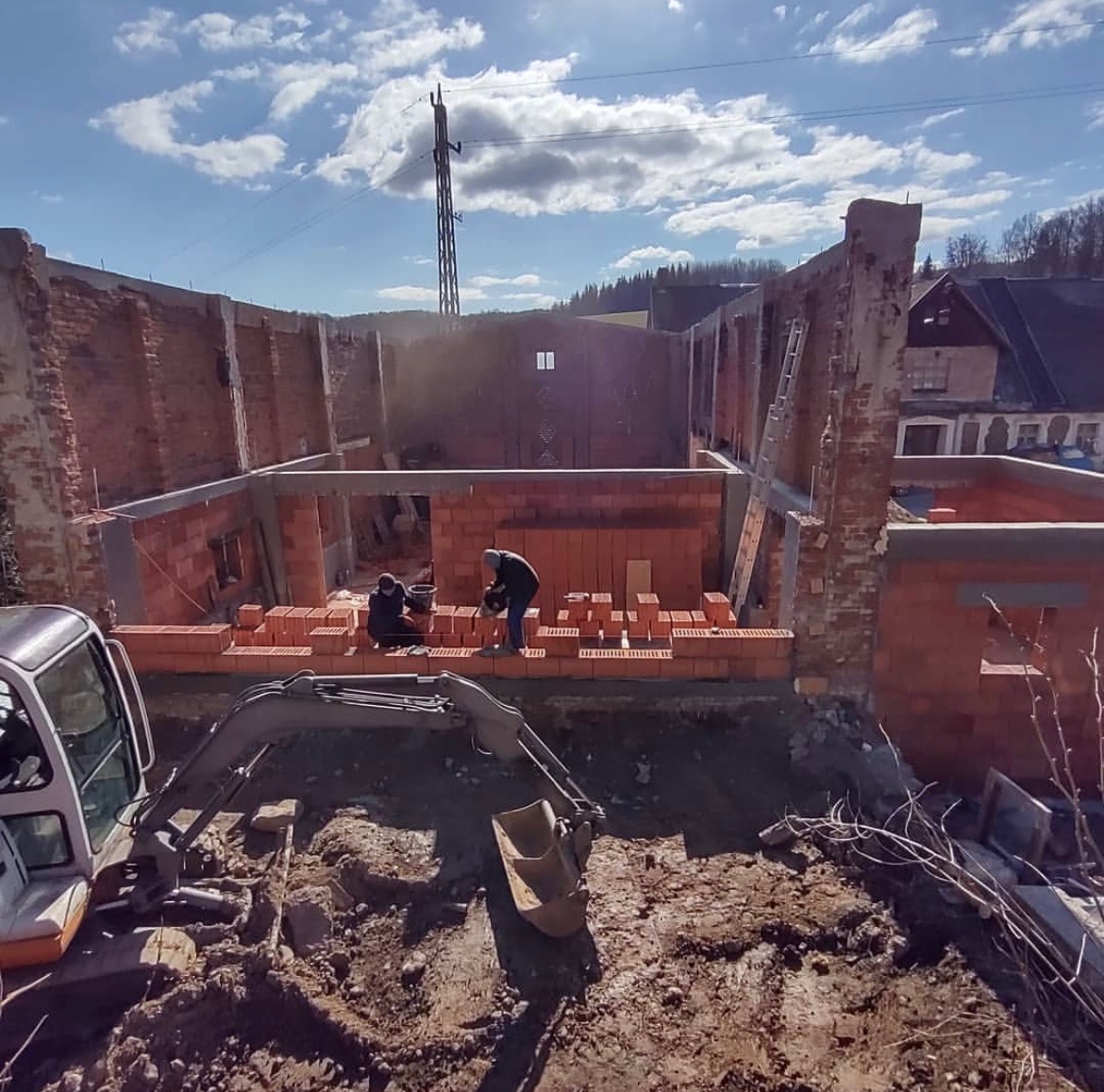 Budowa domu z czerwonej cegły, widoczny fragment koparki, dwóch pracowników murujących ściany, błękitne niebo z chmurami w tle. Prace murarskie w toku.