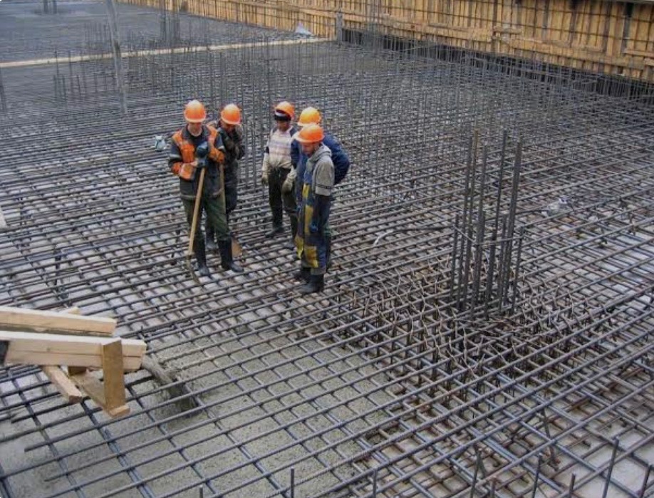 Zbrojenie fundamentów z widocznymi pracownikami w kaskach, przygotowane do zalania betonem. Widoczne szalunki i siatka zbrojeniowa.