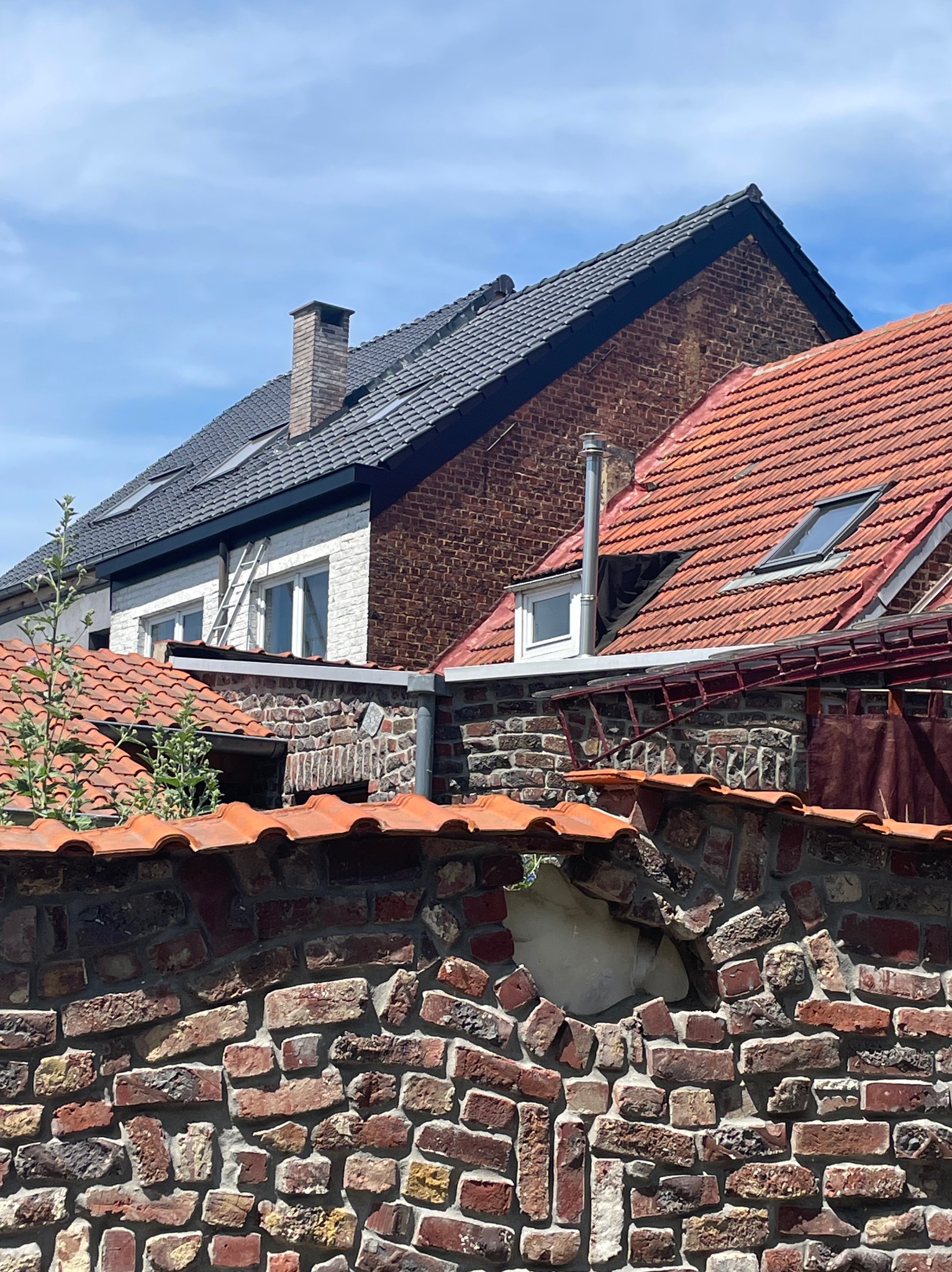 Dwa budynki z cegły o różnych dachach: jeden z czarną dachówką i kominem, drugi z czerwoną dachówką i oknem dachowym. Mur z czerwonej cegły na pierwszym planie.