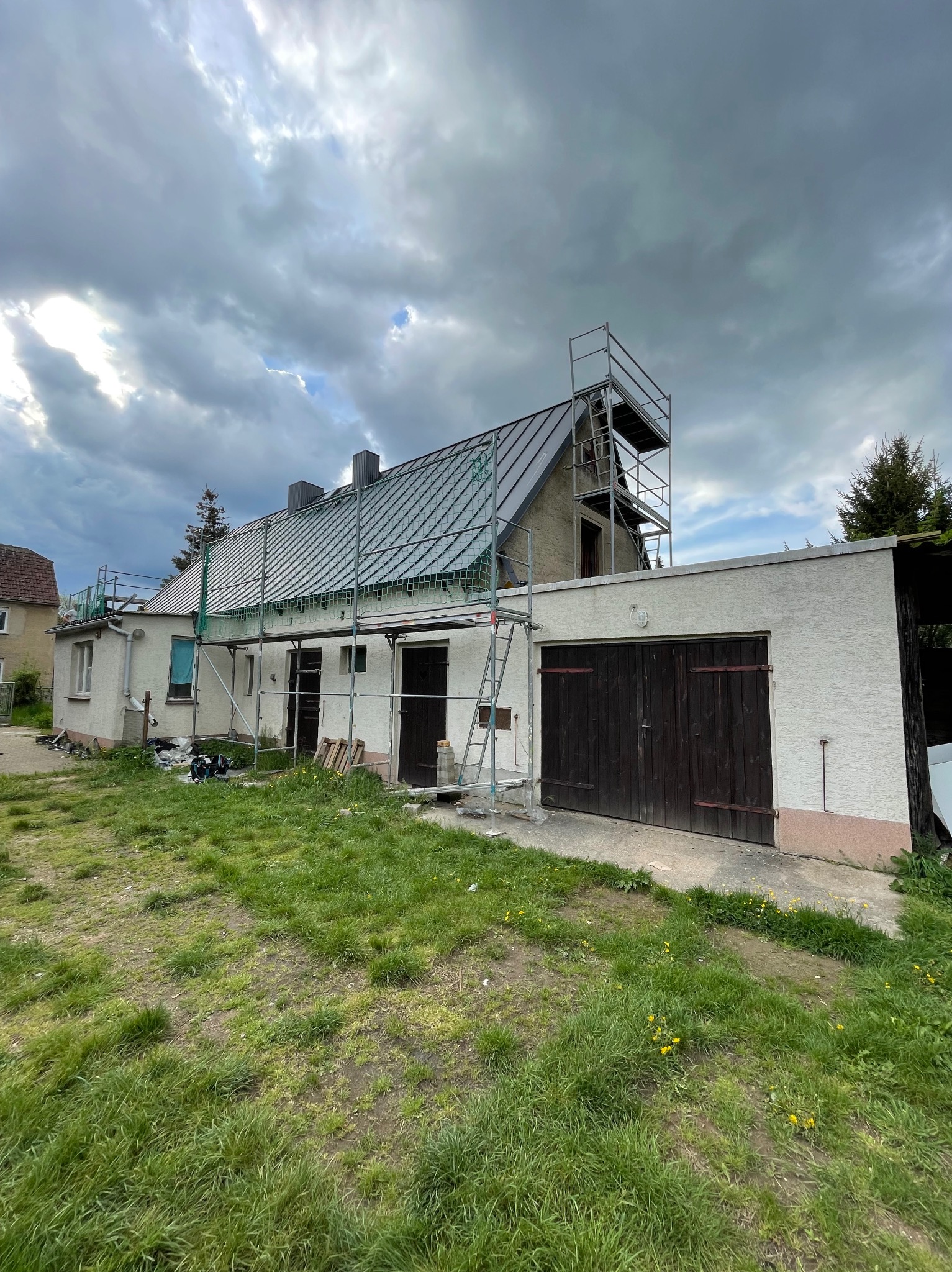 Dom z nowym, metalowym dachem w trakcie montażu rusztowania. Widoczne elementy zabezpieczające. Pochmurne niebo w tle. Ujęcie z poziomu trawnika.