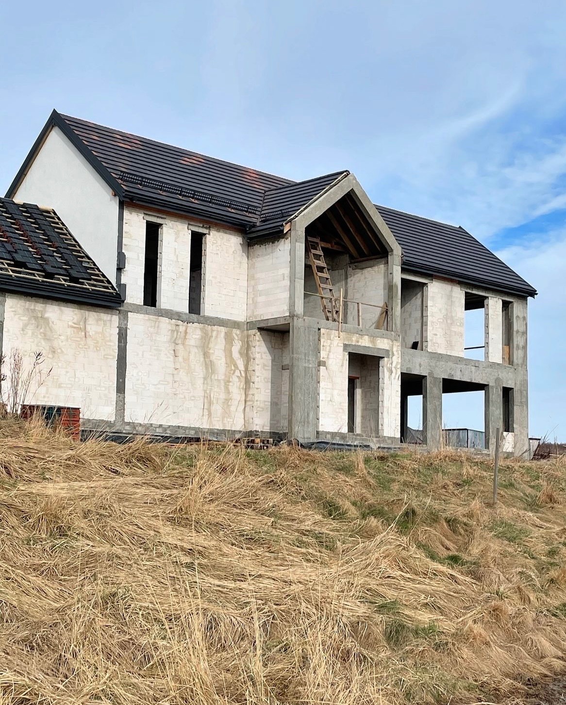 Surowy dom z betonu komórkowego w trakcie budowy, z częściowo ułożonym ciemnym dachem i drewnianą drabiną opartą o ścianę, na tle suchej trawy i błękitnego nieba.