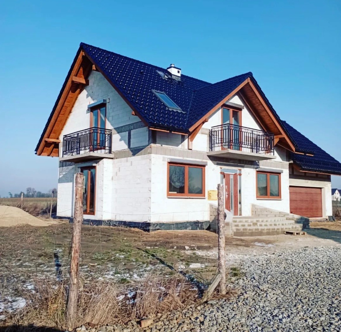 Niedokończony dom z białego bloczka z ciemnym dachem i drewnianymi elementami wykończeniowymi, balkony z czarną balustradą, w tle błękitne niebo.