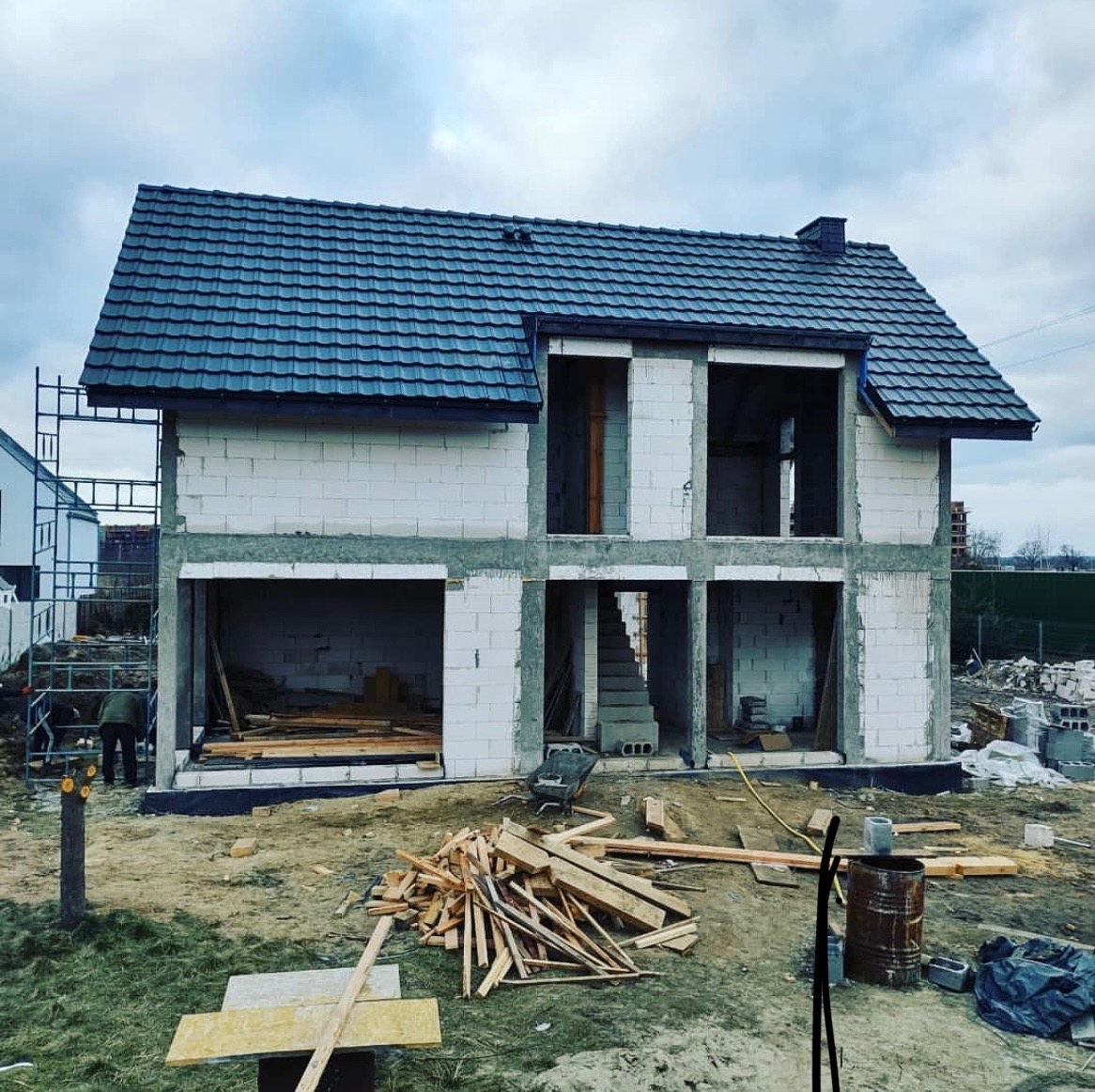 Surowy stan dwukondygnacyjnego domu z szarej cegły z ciemnym dachem, widoczne schody wewnętrzne, drewniane elementy konstrukcyjne i rusztowania.