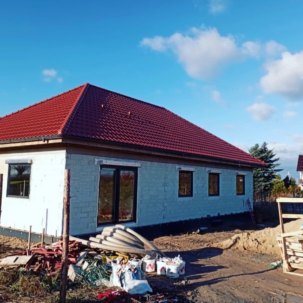 Niski dom w trakcie budowy z czerwoną dachówką, widoczne okna i materiały budowlane na ziemi. Błękitne niebo z chmurami w tle.