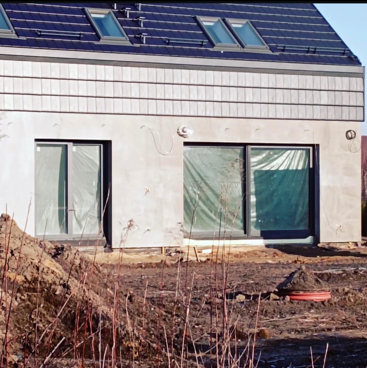 Surowy dom z szarym dachem i oknami dachowymi, częściowo otynkowany, z widocznym wykopem i instalacjami na zewnątrz. Okna zabezpieczone folią.