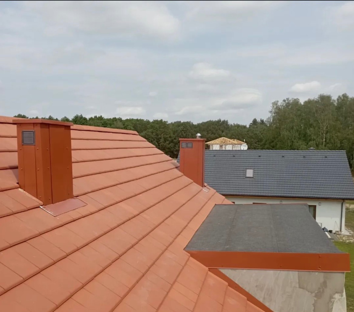 Czerwona dachówka ceramiczna na skośnym dachu domu jednorodzinnego z kominem w kolorze dachówki. W tle widoczny drugi dom z szarą dachówką.