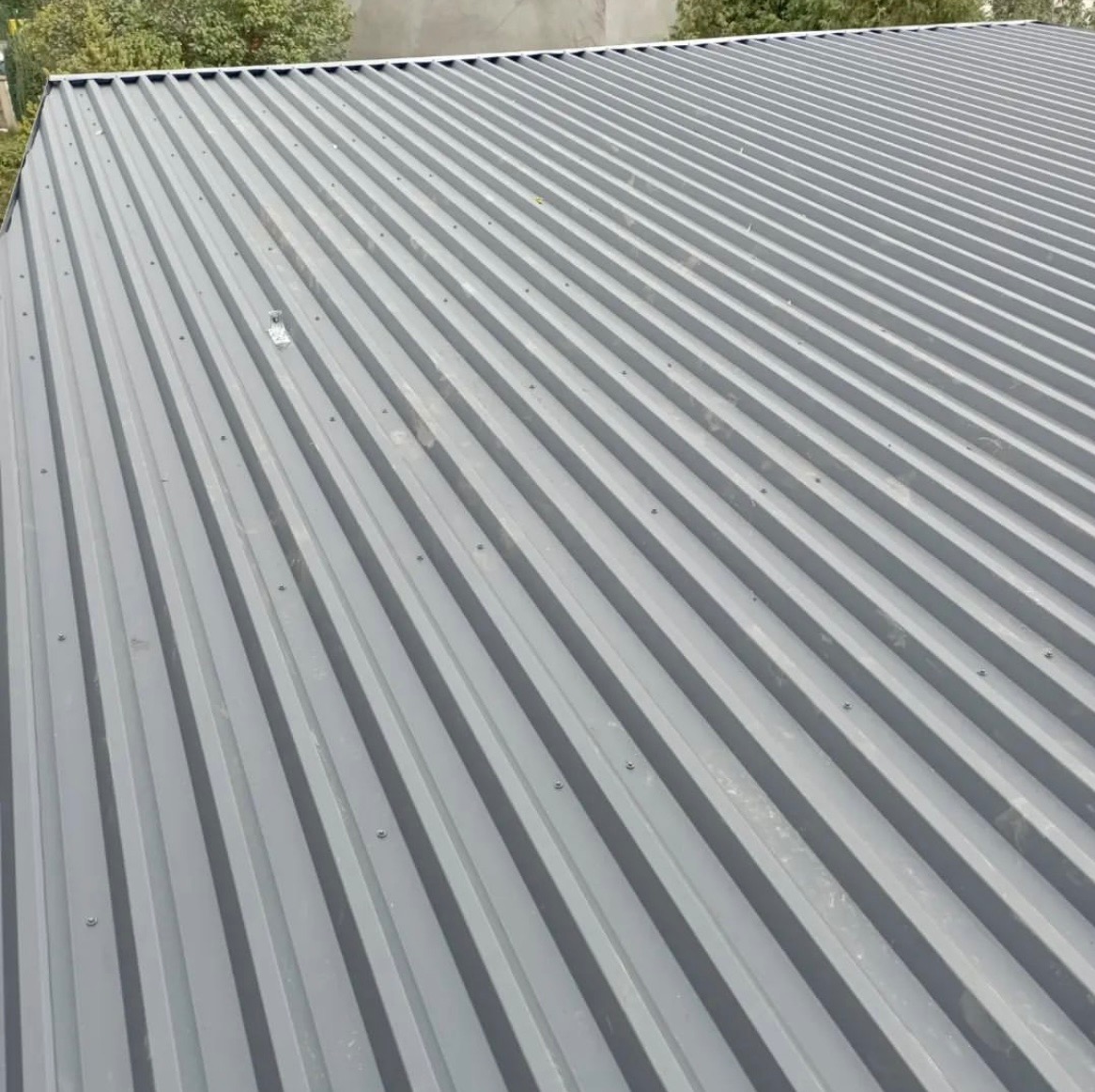Szary dach z blachy trapezowej, widok z góry, równoległe fale, mocowania śrubowe, fragment budynku w tle, drzewa. Nowe pokrycie dachowe.