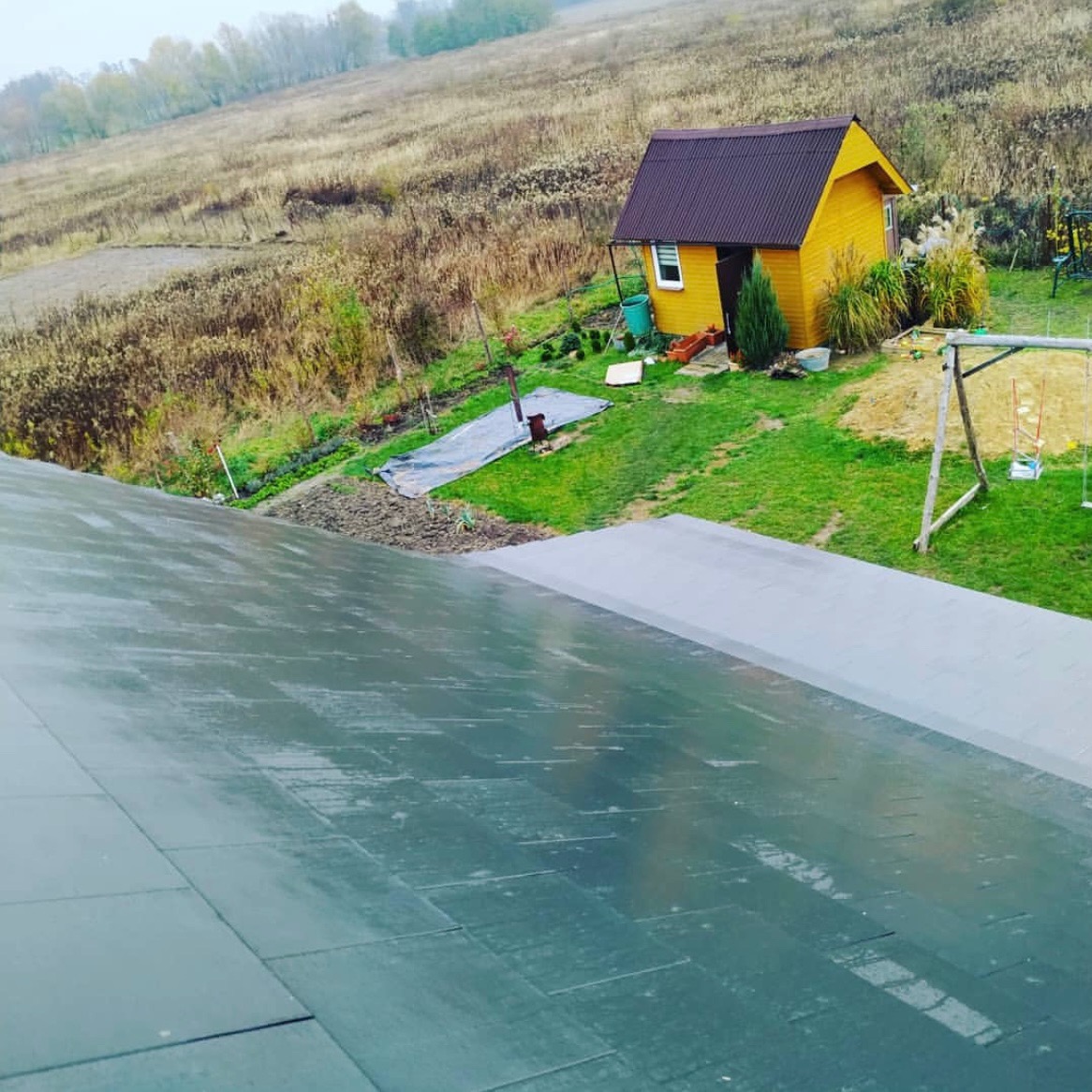 Nowe, grafitowe pokrycie dachowe z papy termozgrzewalnej na tle krajobrazu z małym, żółtym domkiem i huśtawką. Widok z góry, lekko zamglone powietrze.