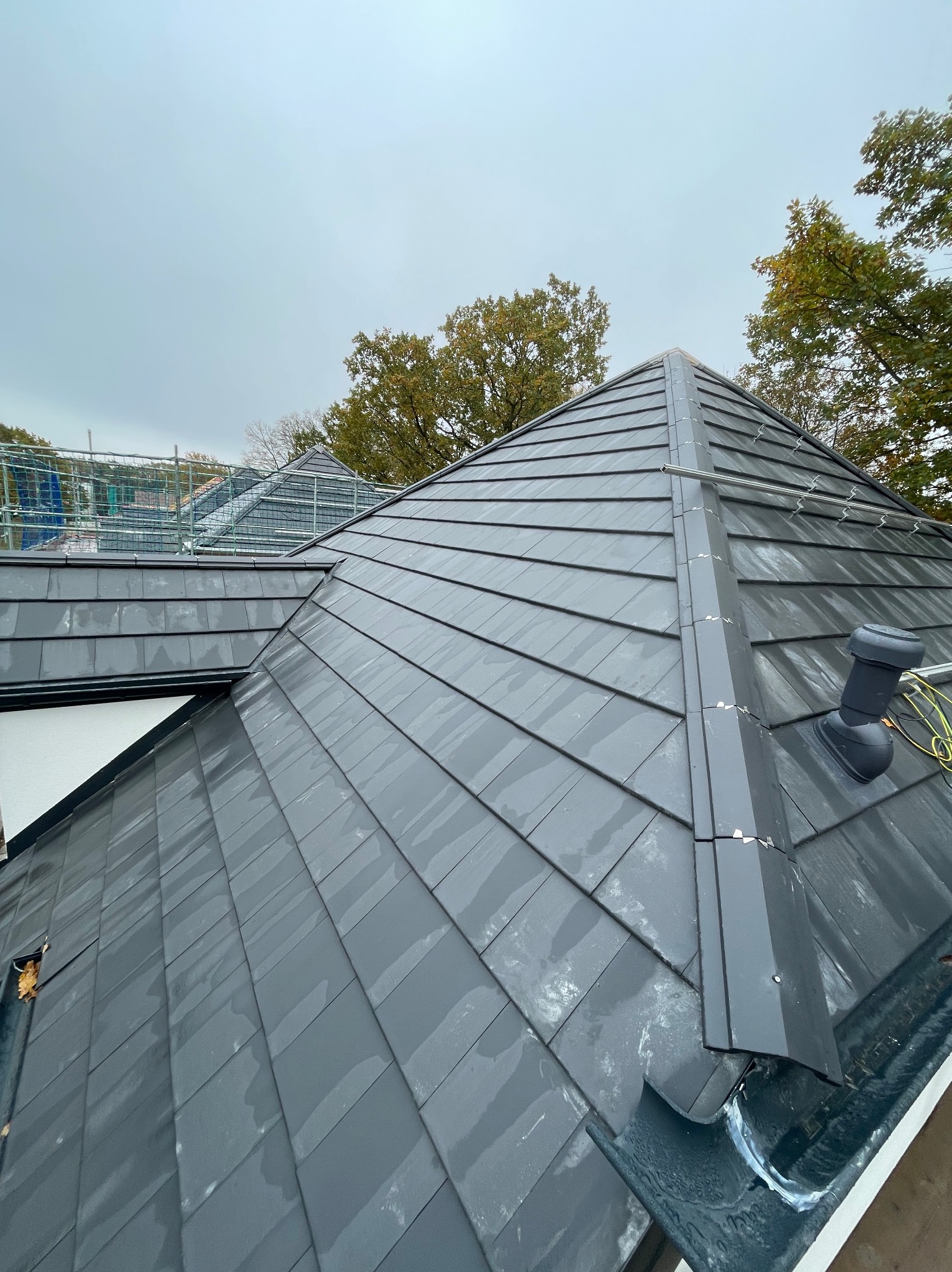 Grafitowy dach z dachówki ceramicznej, widok z góry, z elementami orynnowania i kominkiem wentylacyjnym. W tle drzewa i rusztowania.