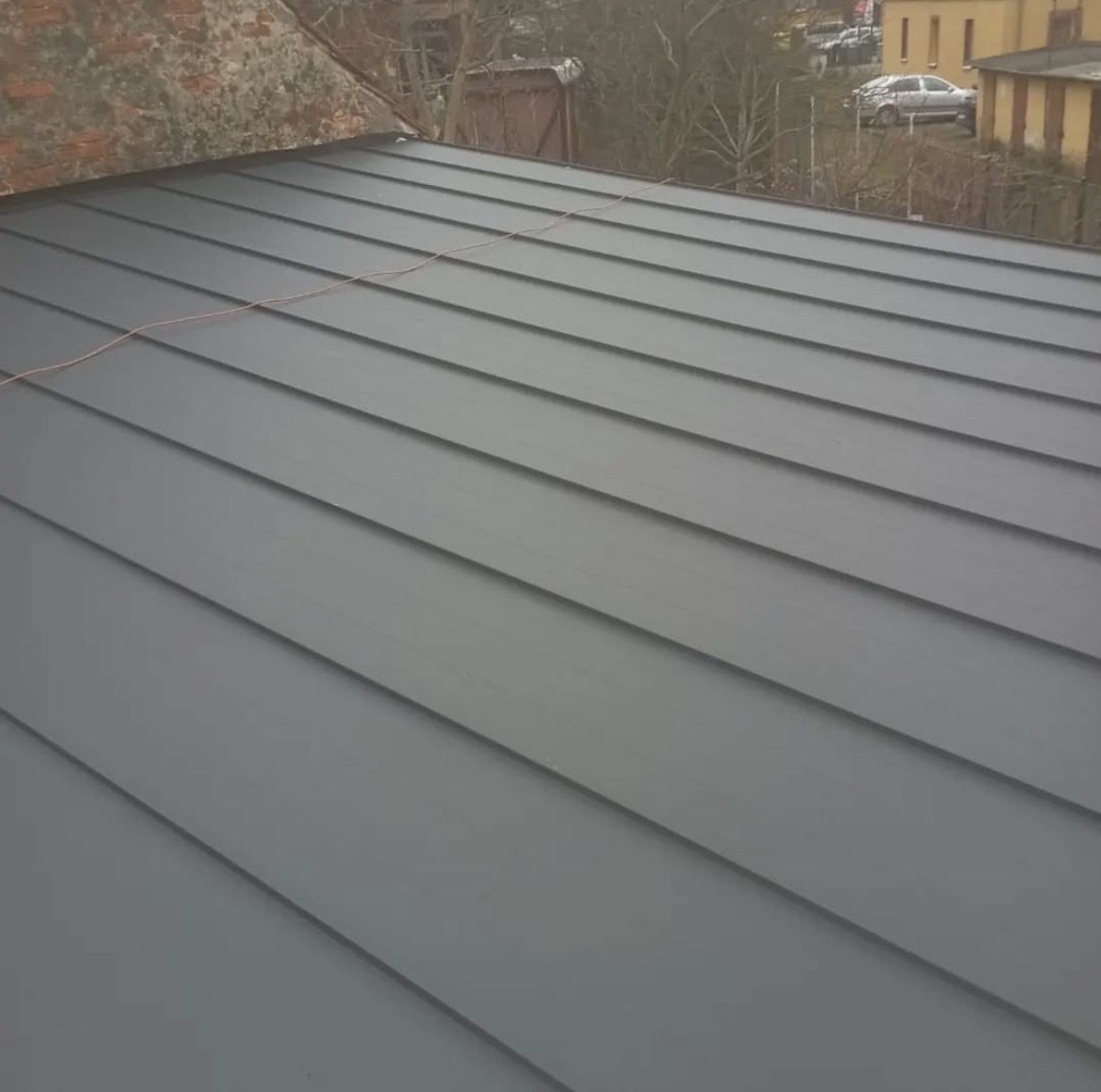 Szary dach z blachy na budynku mieszkalnym, widok z góry. Widoczne równoległe pasy blachy i fragment otoczenia z budynkami w tle.