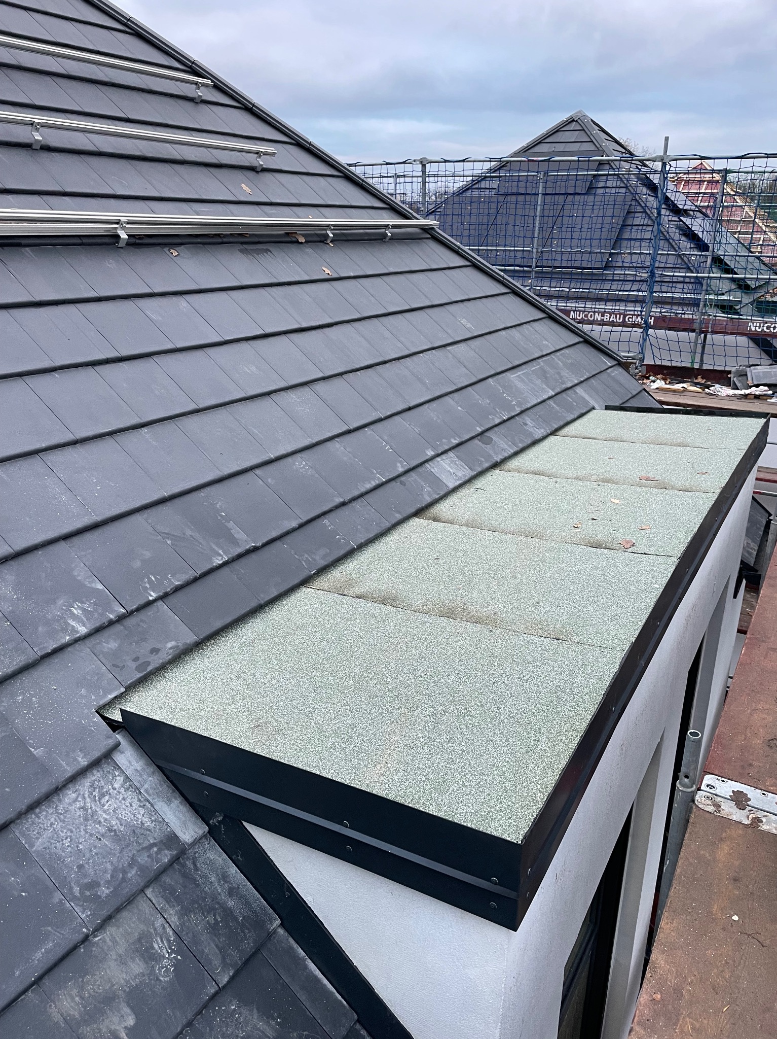 Szary dach z dachówki ceramicznej z elementami montażu paneli solarnych i fragmentem dachu płaskiego pokrytego papą. Widoczne obróbki blacharskie.