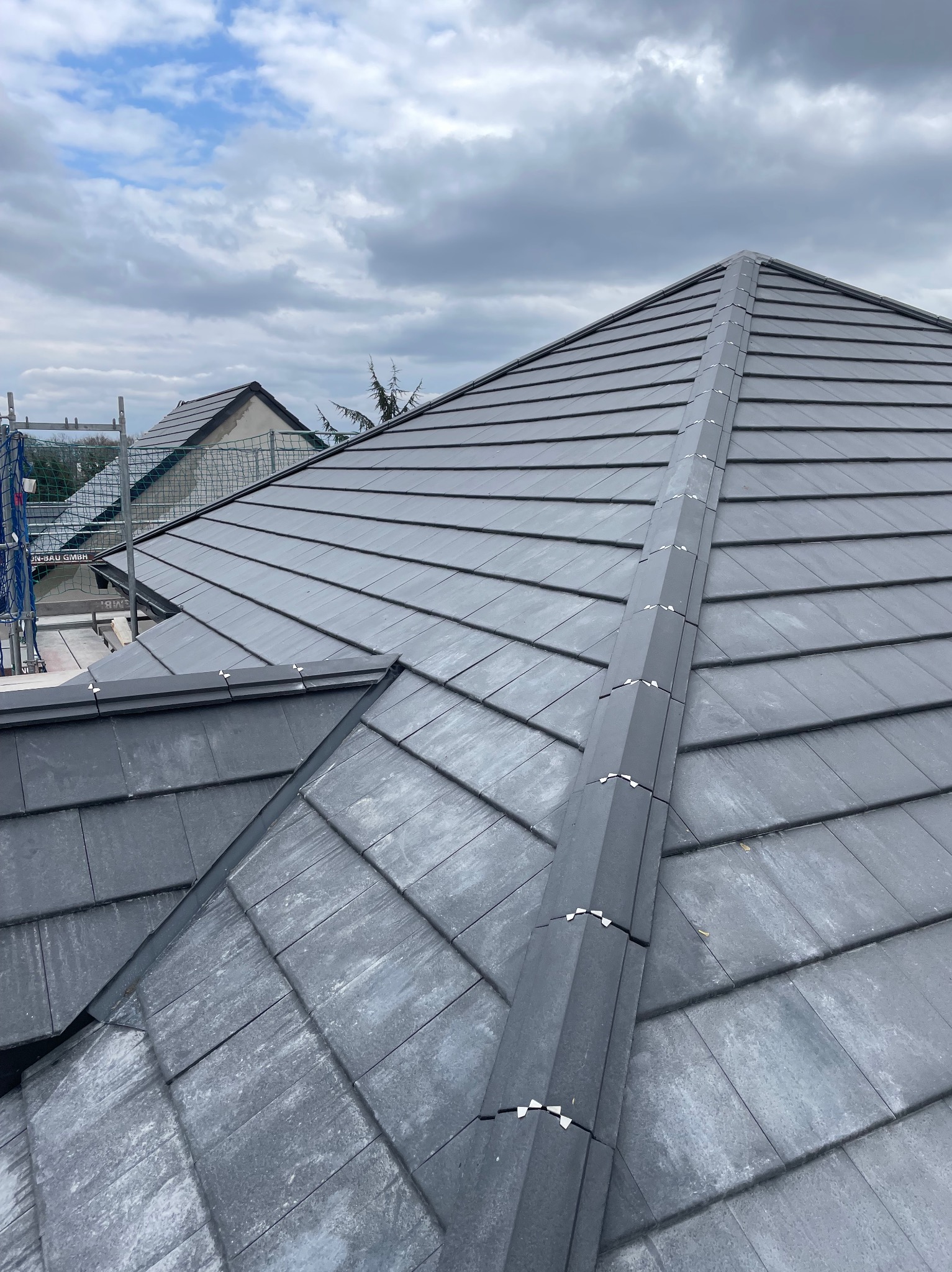 Szary dach z dachówki ceramicznej, widok z góry na kalenicę z mocowaniami, w tle fragment rusztowania i dach sąsiedniego budynku pod pochmurnym niebem.