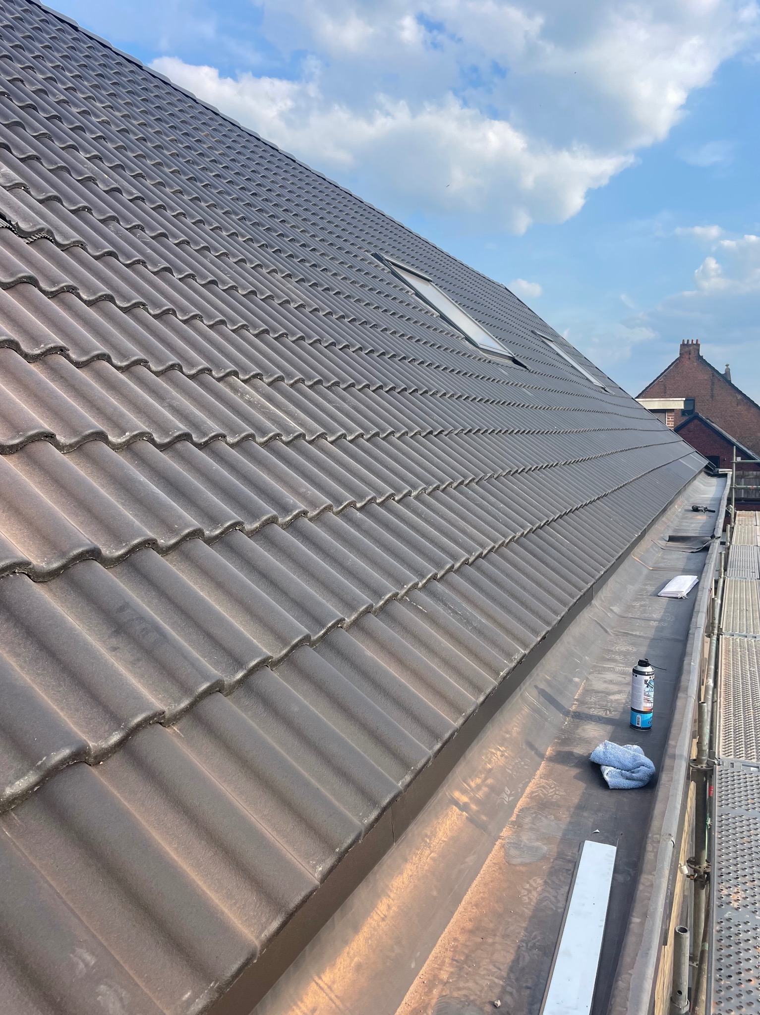 Ciemna dachówka ceramiczna na skośnym dachu z oknami połaciowymi, widok z rusztowania. Obróbki blacharskie wzdłuż krawędzi dachu. Na pierwszym planie narzędzia.