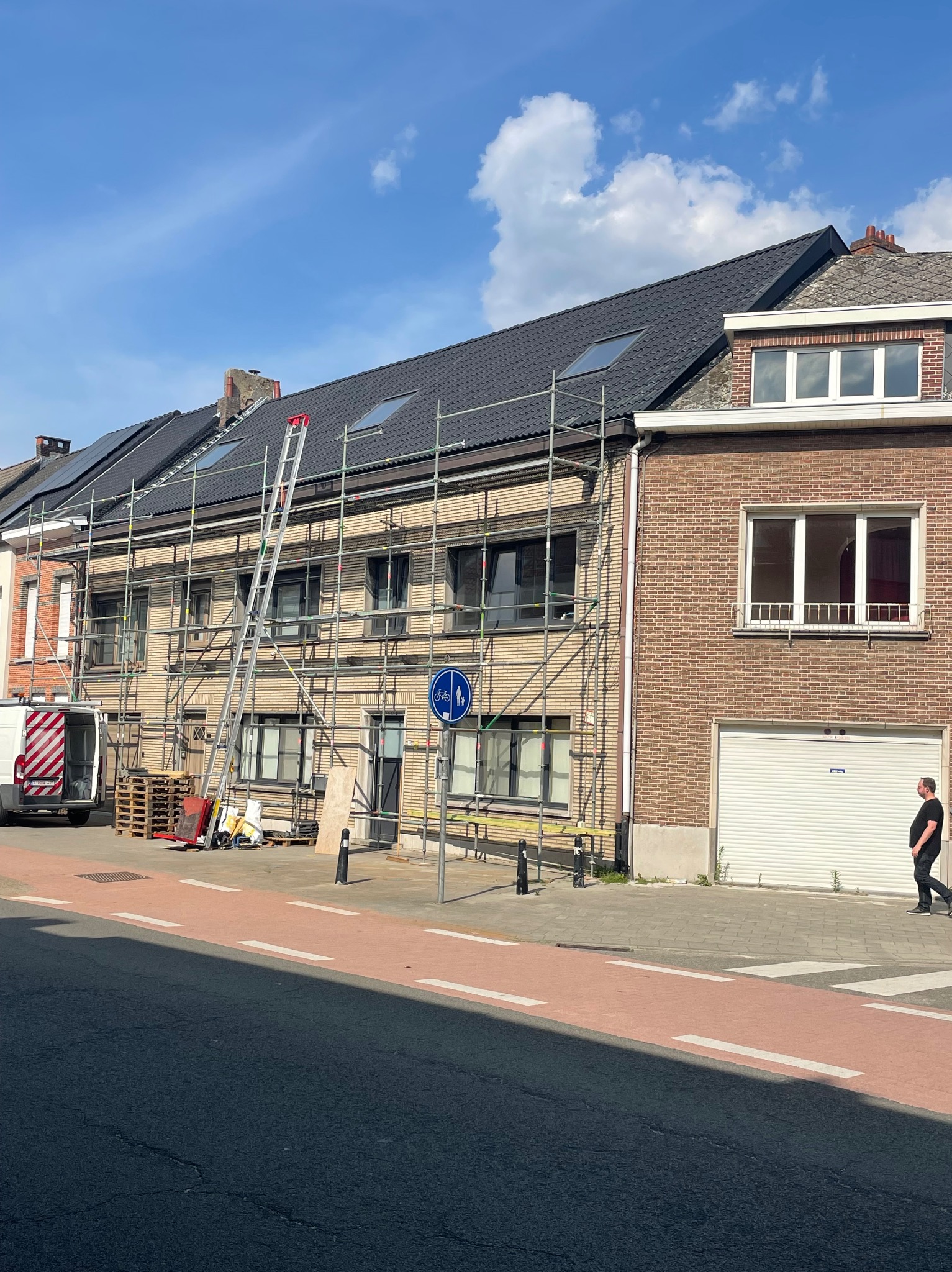 Dom w rusztowaniach z nowym, ciemnym dachem i oknami dachowymi. Na chodniku przechodzień, a obok zaparkowany van. Słoneczny dzień.