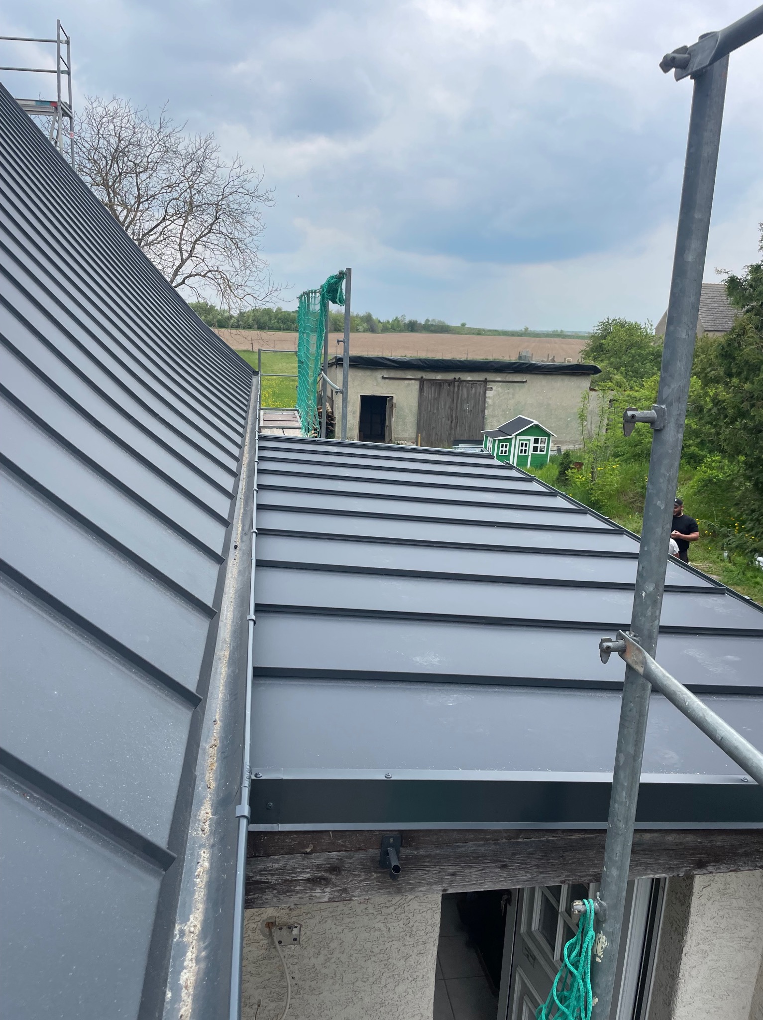 Szary dach z blachy na rąbek stojący, widok z rusztowania. W tle budynek gospodarczy i zielony domek dla dzieci. Nowoczesne krycie dachu.