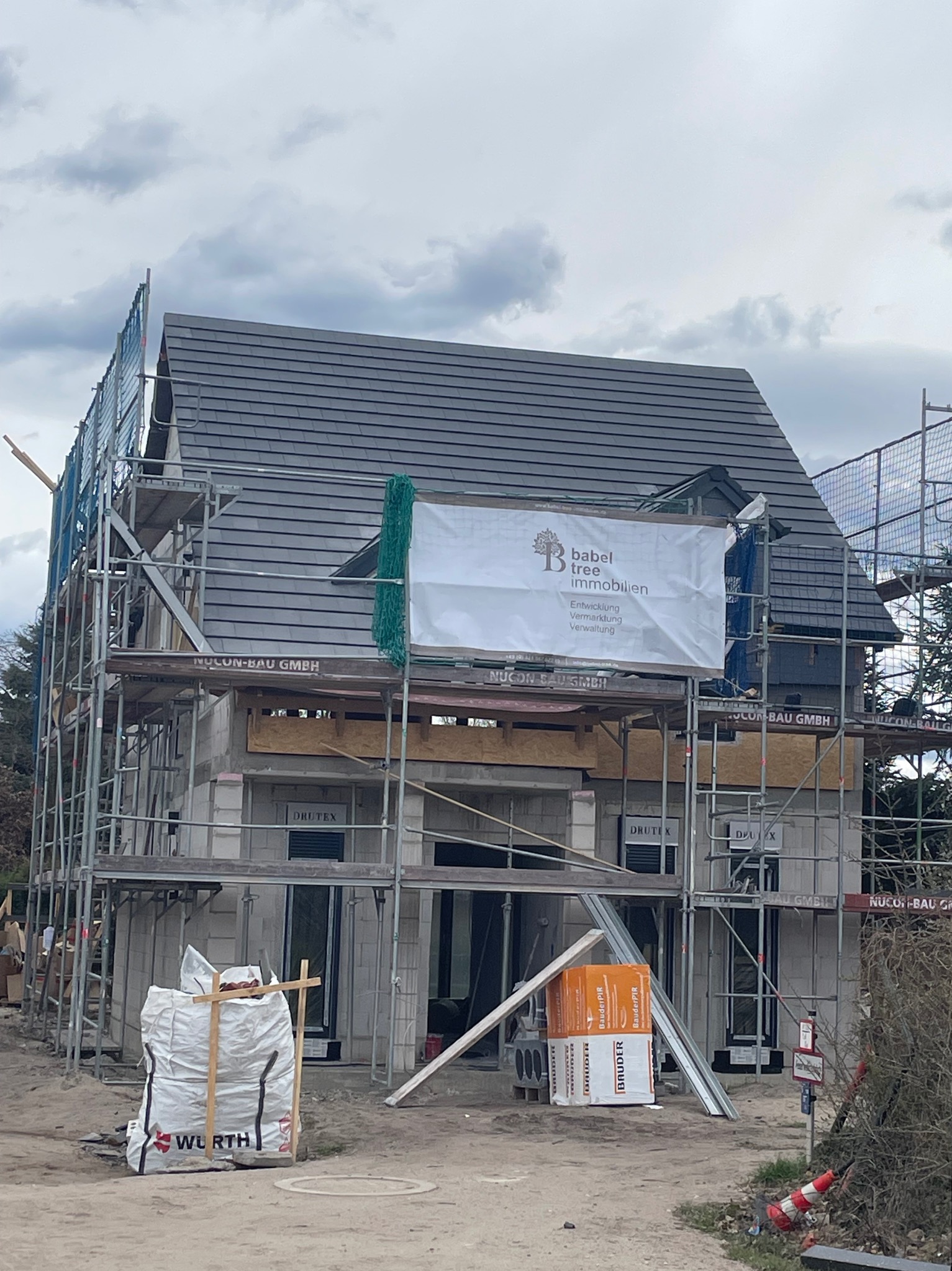 Dom w trakcie budowy z szarym dachem, otoczony rusztowaniami i materiałami budowlanymi. Na rusztowaniu baner reklamowy. Widoczne okna DRUTEX.