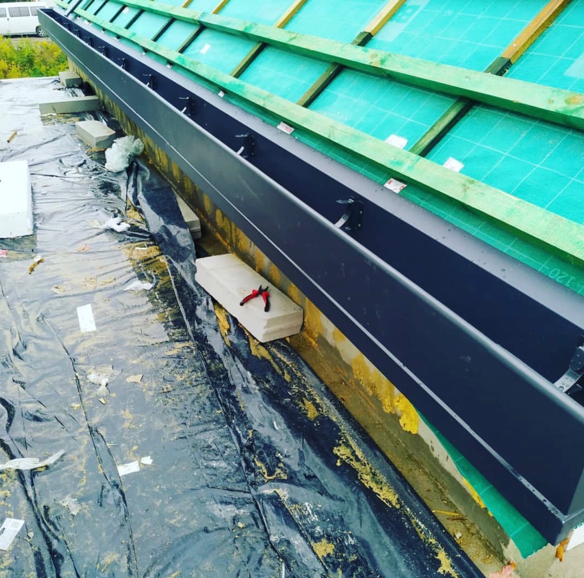 Montaż czarnej rynny dachowej na zielonej membranie, widoczne narzędzia i elementy konstrukcji dachu. Ujęcie z góry, podkreślające detale instalacji.