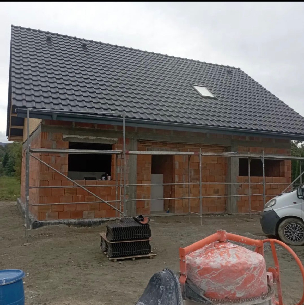 Budowa domu w trakcie: murowane ściany z pustaków, nowy dach z ciemnej dachówki, rusztowania. Na pierwszym planie betoniarka i paleta z dachówką.
