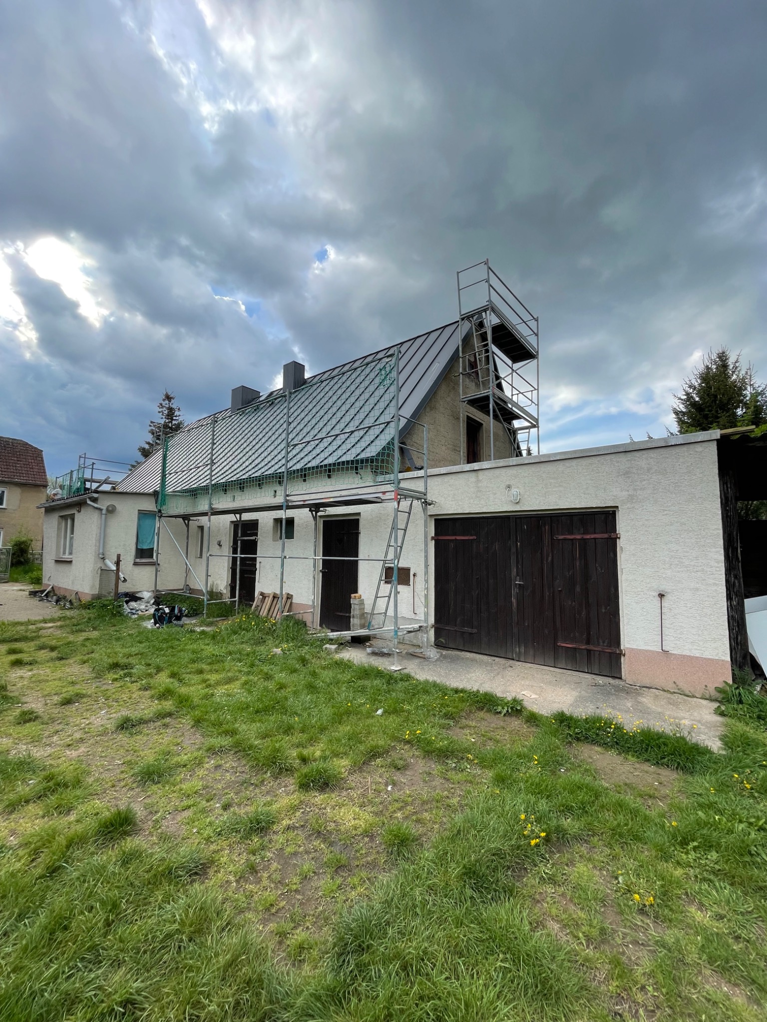 Dom jednorodzinny w trakcie prac dekarskich, widoczne rusztowania i zabezpieczenia na dachu. Pochmurne niebo w tle.