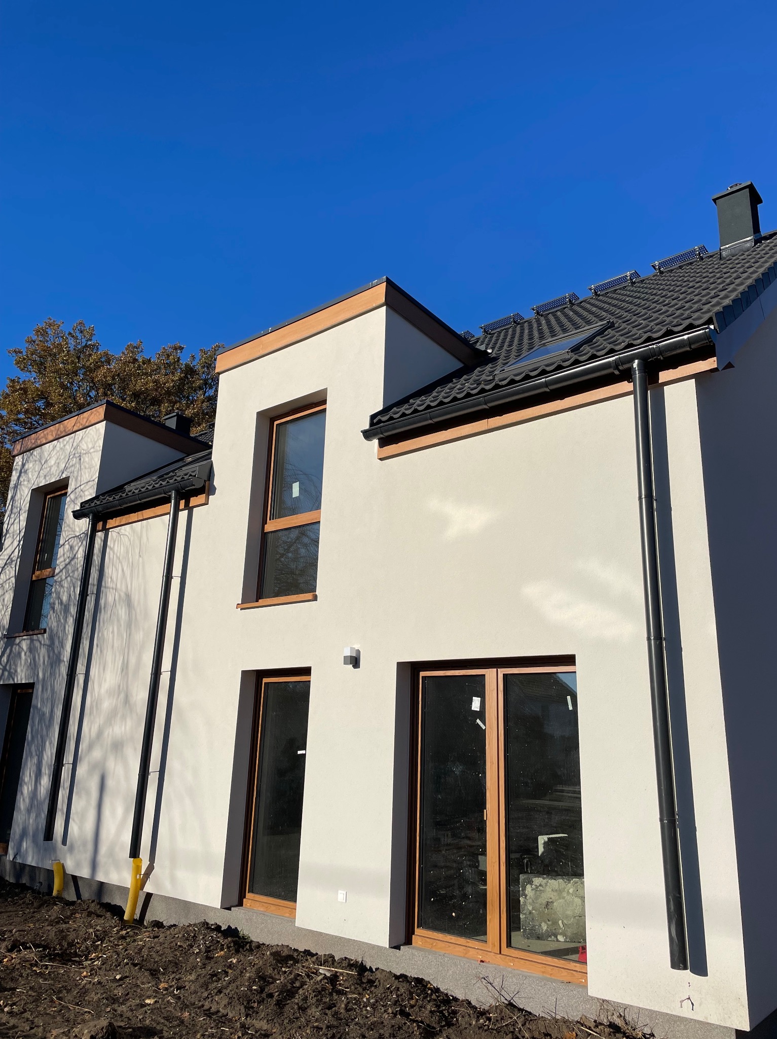 Nowoczesny dom z ciemną dachówką i panelami solarnymi, jasną elewacją, drewnianymi ramami okiennymi i czarnymi rynnami w słoneczny dzień.