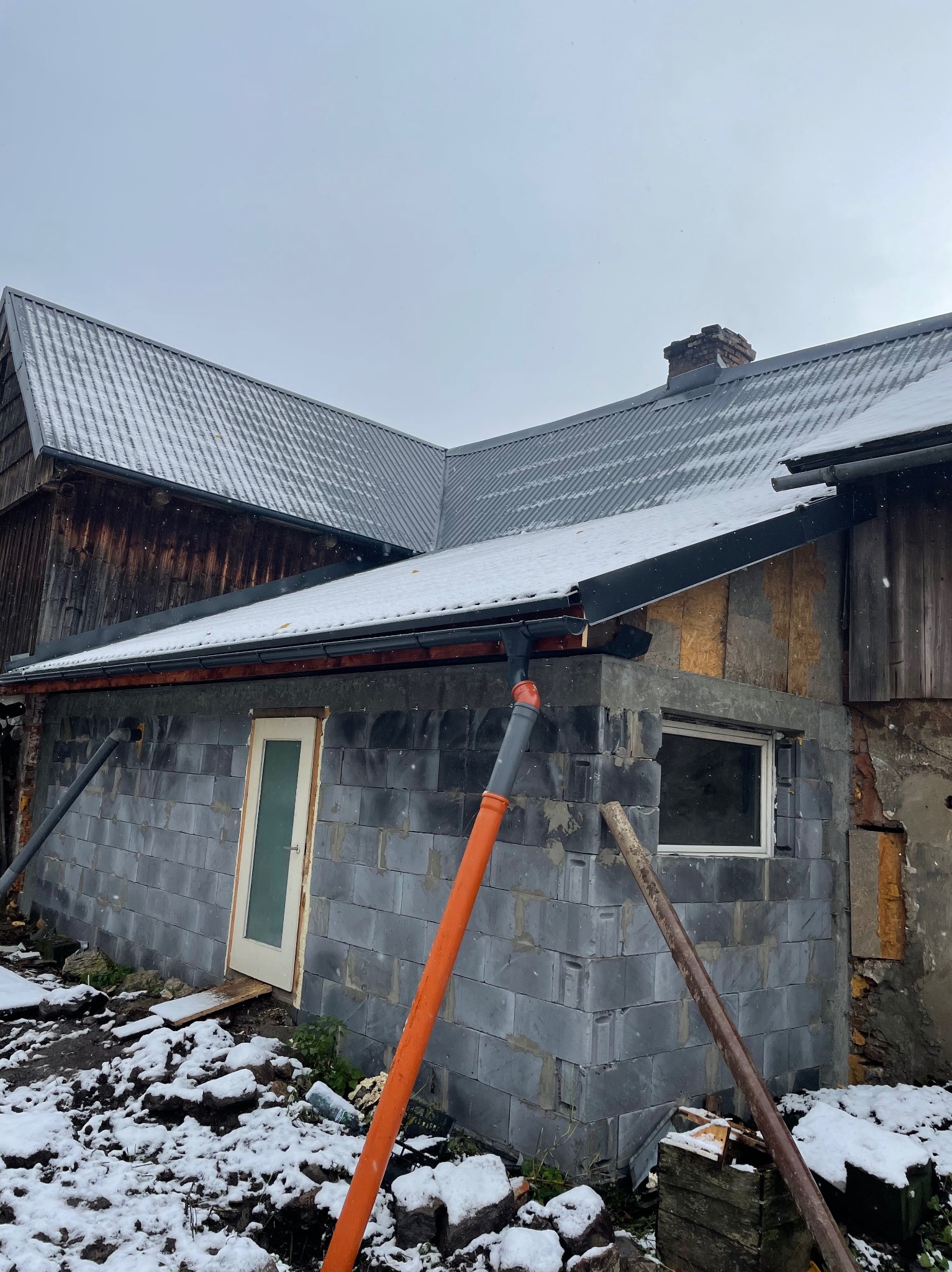 Budynek w trakcie budowy z szarej cegły, widoczne orynnowanie i fragment dachu pokrytego śniegiem. Częściowo ocieplona ściana i okno. Zima.