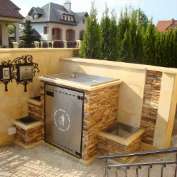 Zabudowa śmietnika z kamienia i tynku w ogrodzie, z metalowymi elementami i ozdobną skrzynką na listy, na tle domów i zieleni.