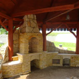 Kremowy, murowany grill z kamienia pod drewnianą altaną z oświetleniem, widok z zewnątrz na ogród z białym płotem i szklarnią.