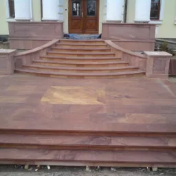 Zewnętrzne schody z czerwonego kamienia prowadzące do wejścia budynku z kolumnami, częściowo ukończone, z widocznymi elementami konstrukcyjnymi w tle.