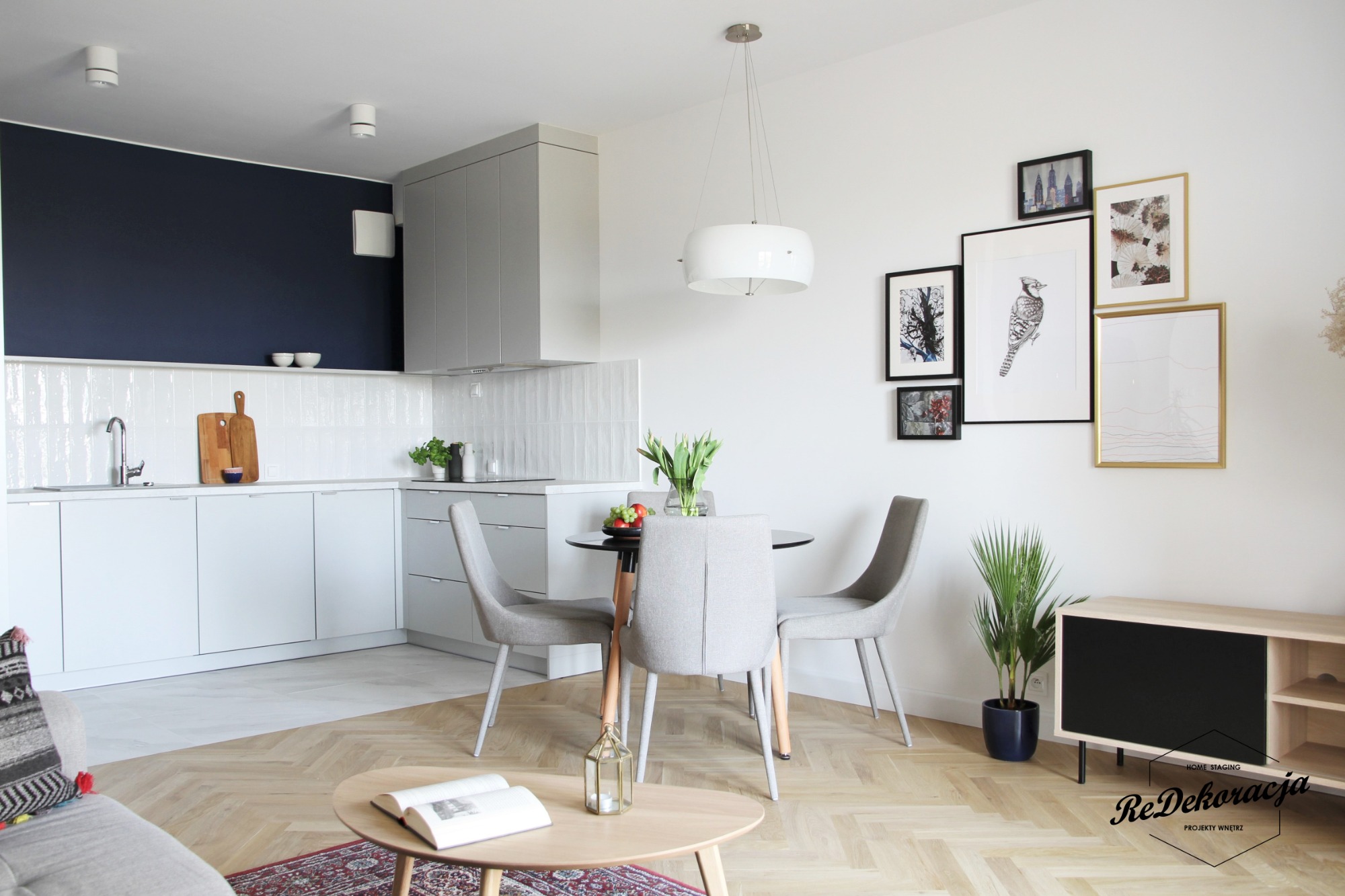 Jasne, nowoczesne wnętrze mieszkania z aneksem kuchennym, okrągłym stołem jadalnym i galerią obrazów na ścianie, oświetlone lampą wiszącą i naturalnym światłem.
