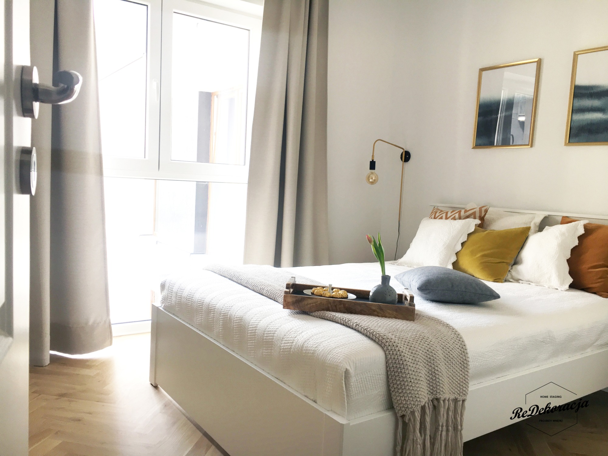 Jasna sypialnia z białym łóżkiem, drewnianą podłogą w jodełkę i dekoracyjnym oświetleniem ściennym, stylizowana z tacą śniadaniową i elementami tekstylnymi, widoczny fragment drzwi z klamką.