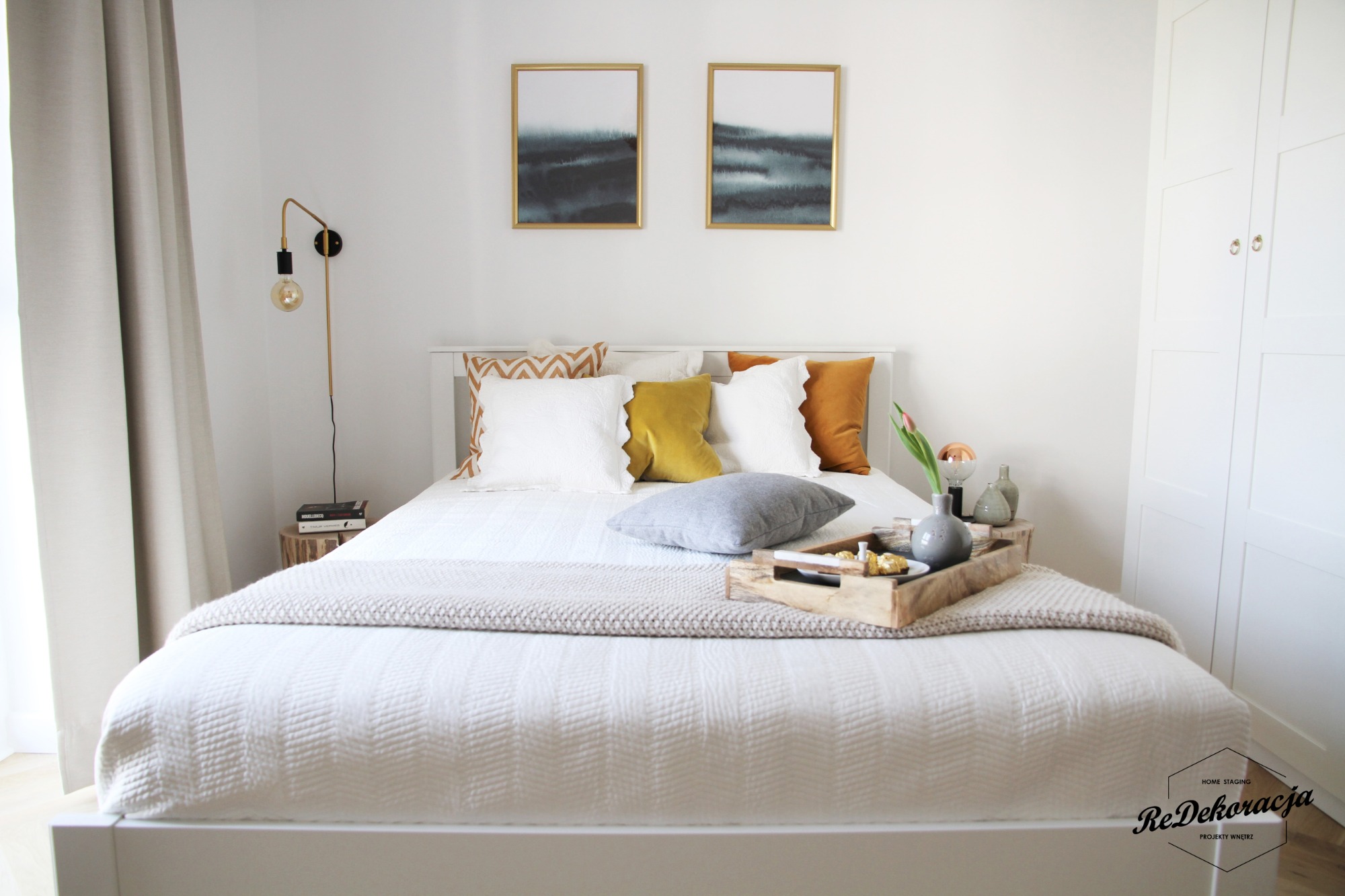 Jasna sypialnia z białym łóżkiem, poduszkami w odcieniach bieli, szarości, żółci i pomarańczy, drewnianą tacą z przekąskami, obrazami na ścianie i lampą ścienną.