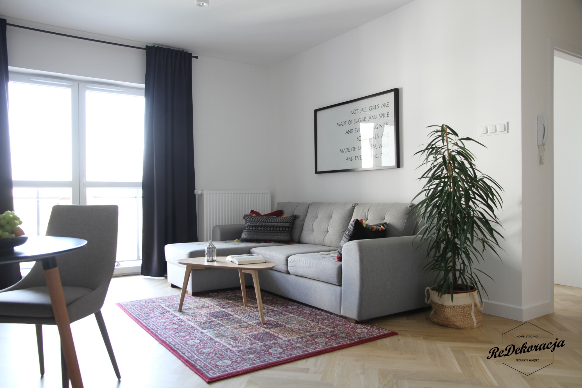 Jasne wnętrze salonu z szarą sofą, drewnianym stolikiem kawowym, wzorzystym dywanem i rośliną doniczkową, uzupełnione czarnymi zasłonami i plakatem z napisem, na podłodze parkiet w jodełkę.