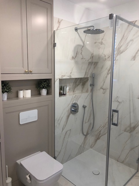 Nowoczesna łazienka z kabiną prysznicową typu walk-in, marmurowymi płytkami i zabudową meblową w odcieniach beżu, z dekoracyjnymi roślinami i akcesoriami toaletowymi w półce.