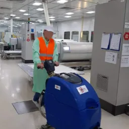 Pracownik w fartuchu i kamizelce odblaskowej obsługuje niebieską maszynę do mycia podłóg w sterylnym pomieszczeniu z panelowym oświetleniem sufitowym i metalowymi szafami sterowniczymi.