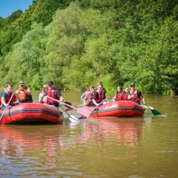TRAVELSAGE SPÓŁKA Z OGRANICZONĄ ODPOWIEDZIALNOŚCIĄ - Dwa czerwone pontony z grupami osób w kamizelkach płyną po rzece, otoczone zielenią drzew. Słoneczny dzień, aktywny wypoczynek na wodzie.